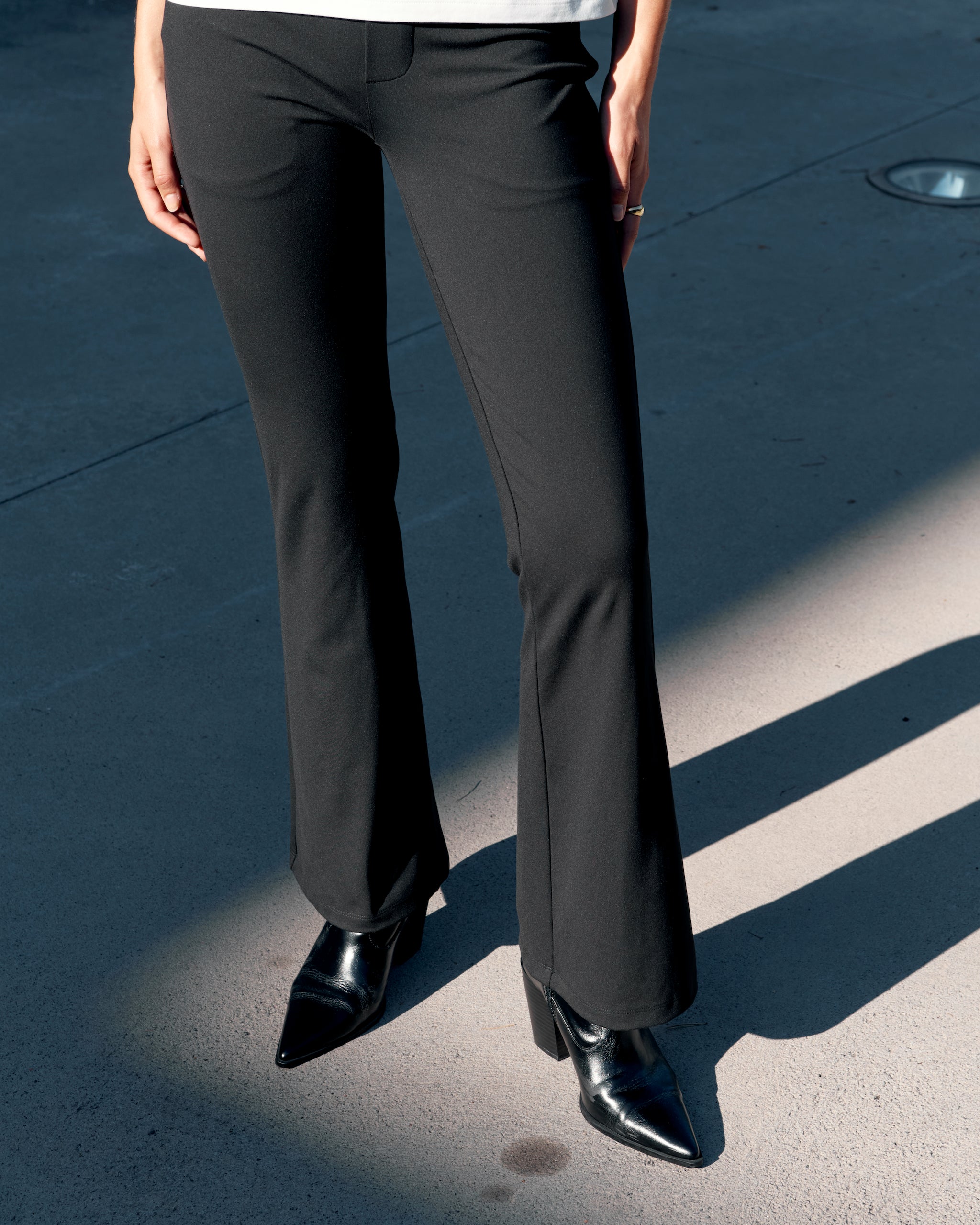 Close-up of black Ninepine asana bootcut office trousers on a model, showing a smooth tailored fit through the legs with black boots in natural light.