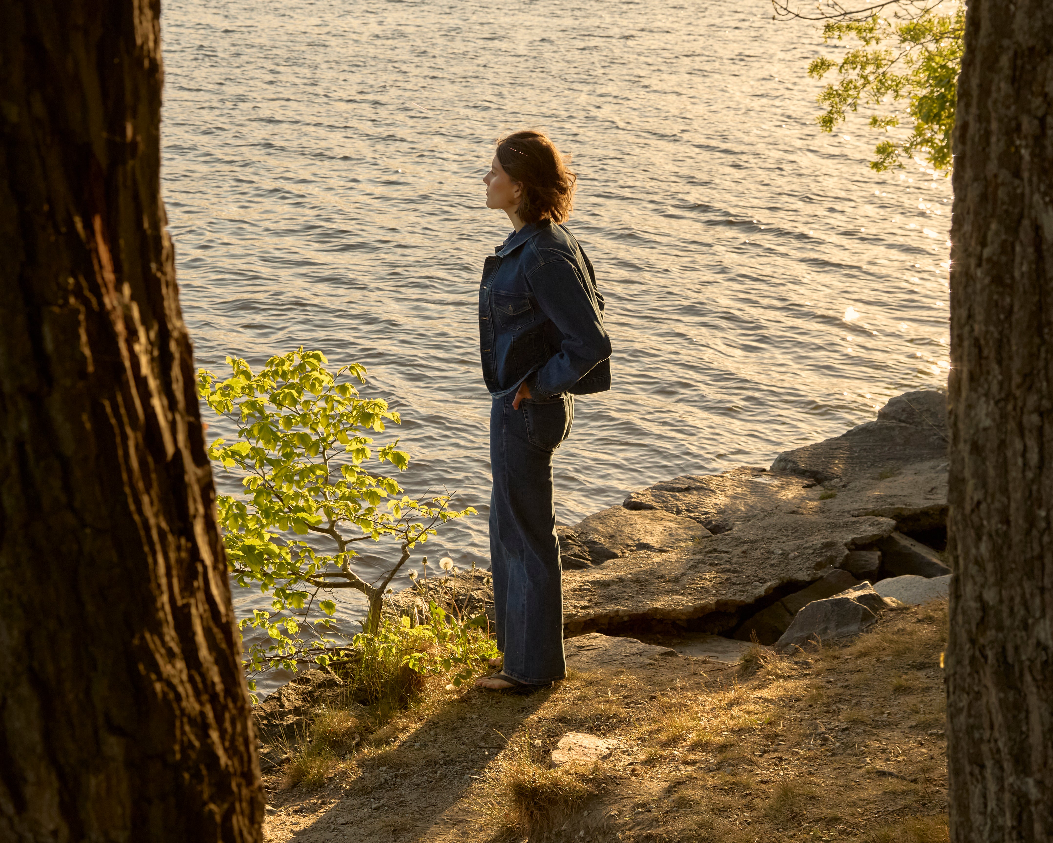 Woman in mid-blue Ninepine ComfortDenim jacket and relaxed jeans stands on a forested Swedish lakeshore at golden hour, gazing over sunlit water.
