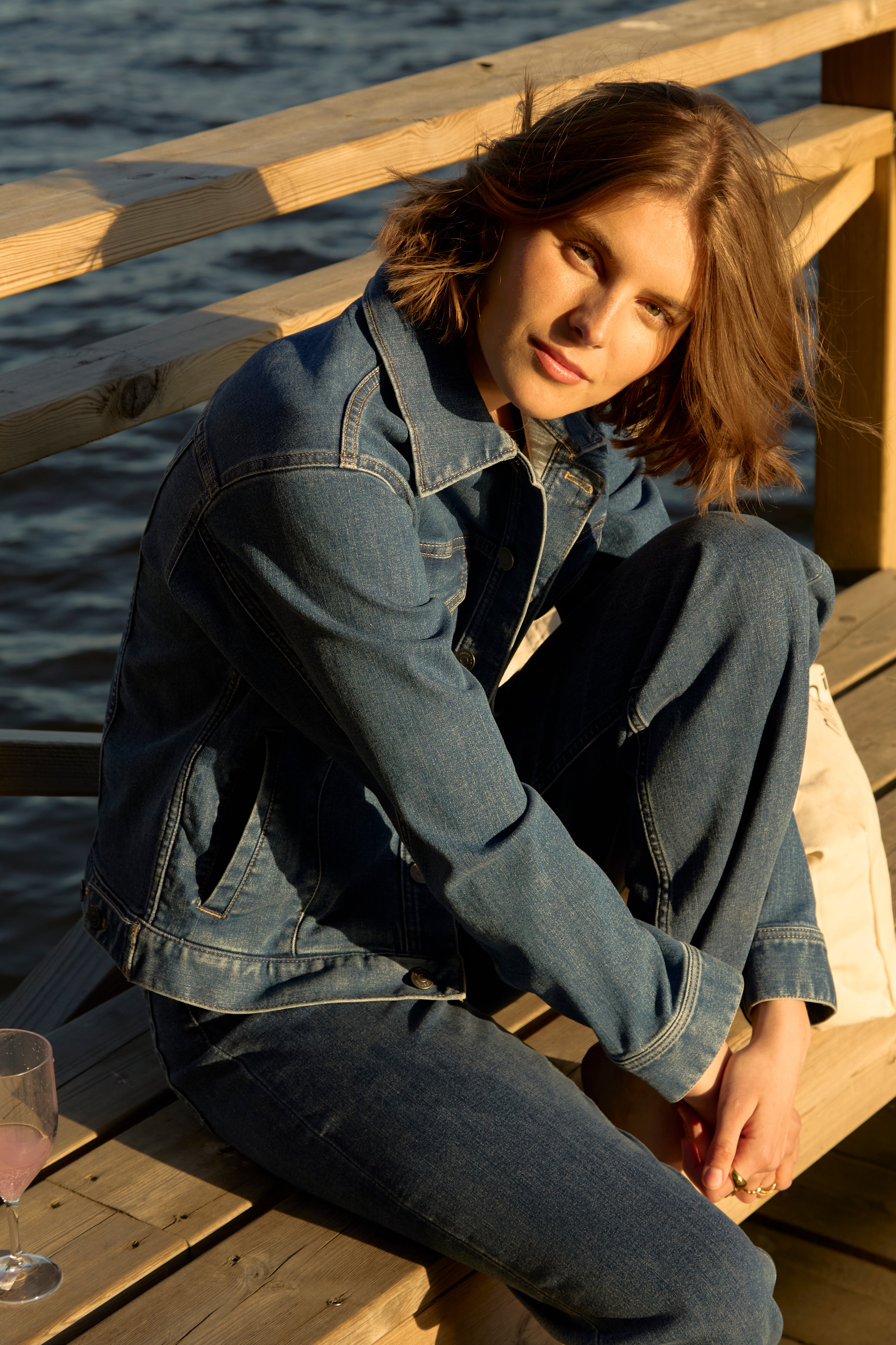 Woman seated on a wooden Swedish lakeside pier at golden hour, dressed in mid-blue Ninepine ComfortDenim jacket and matching jeans, mellow summer evening vibe.