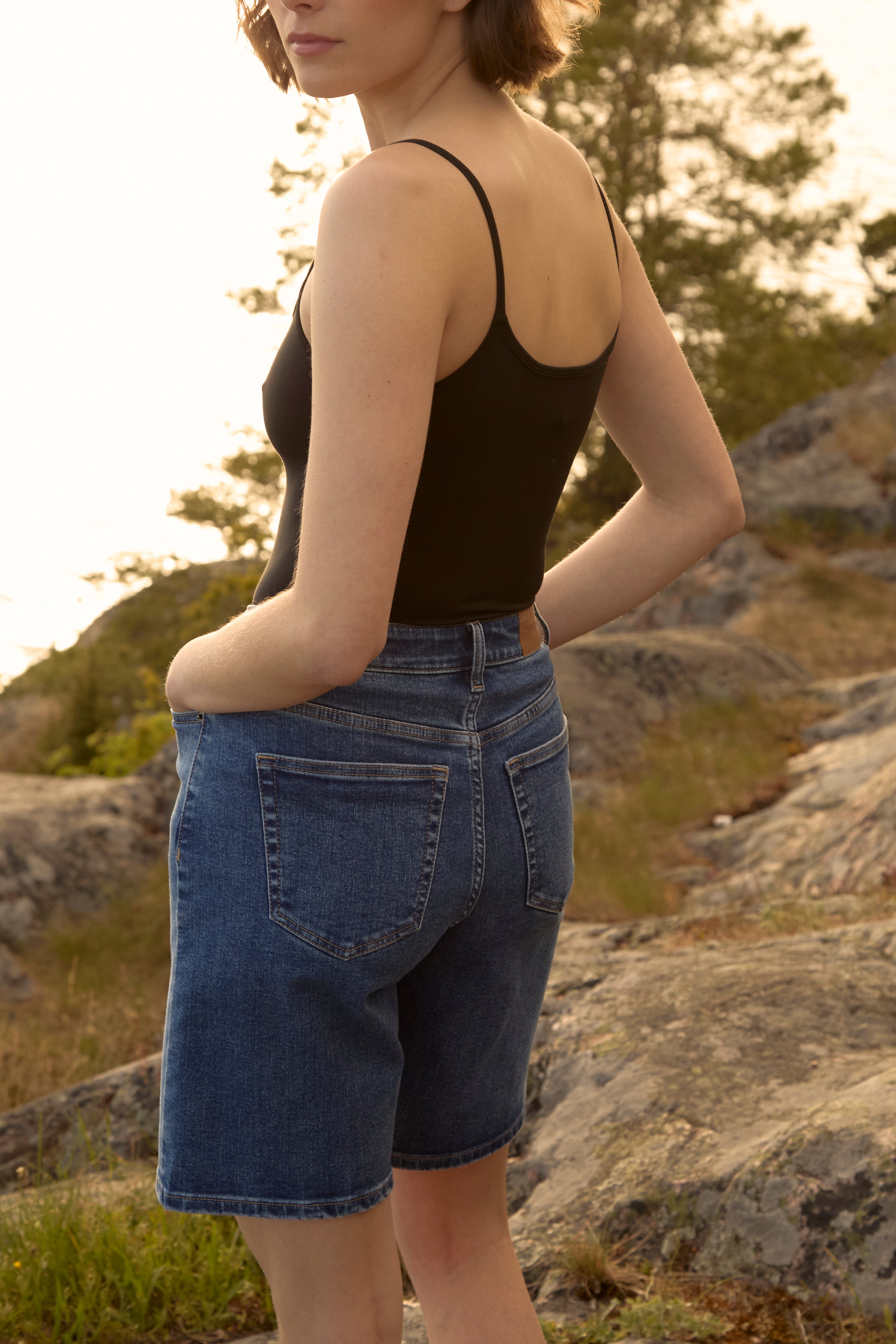 Woman at golden Swedish archipelago sunset wearing mid-blue Ninepine ComfortDenim shorts and black strappy tank, relaxed summer vibe.