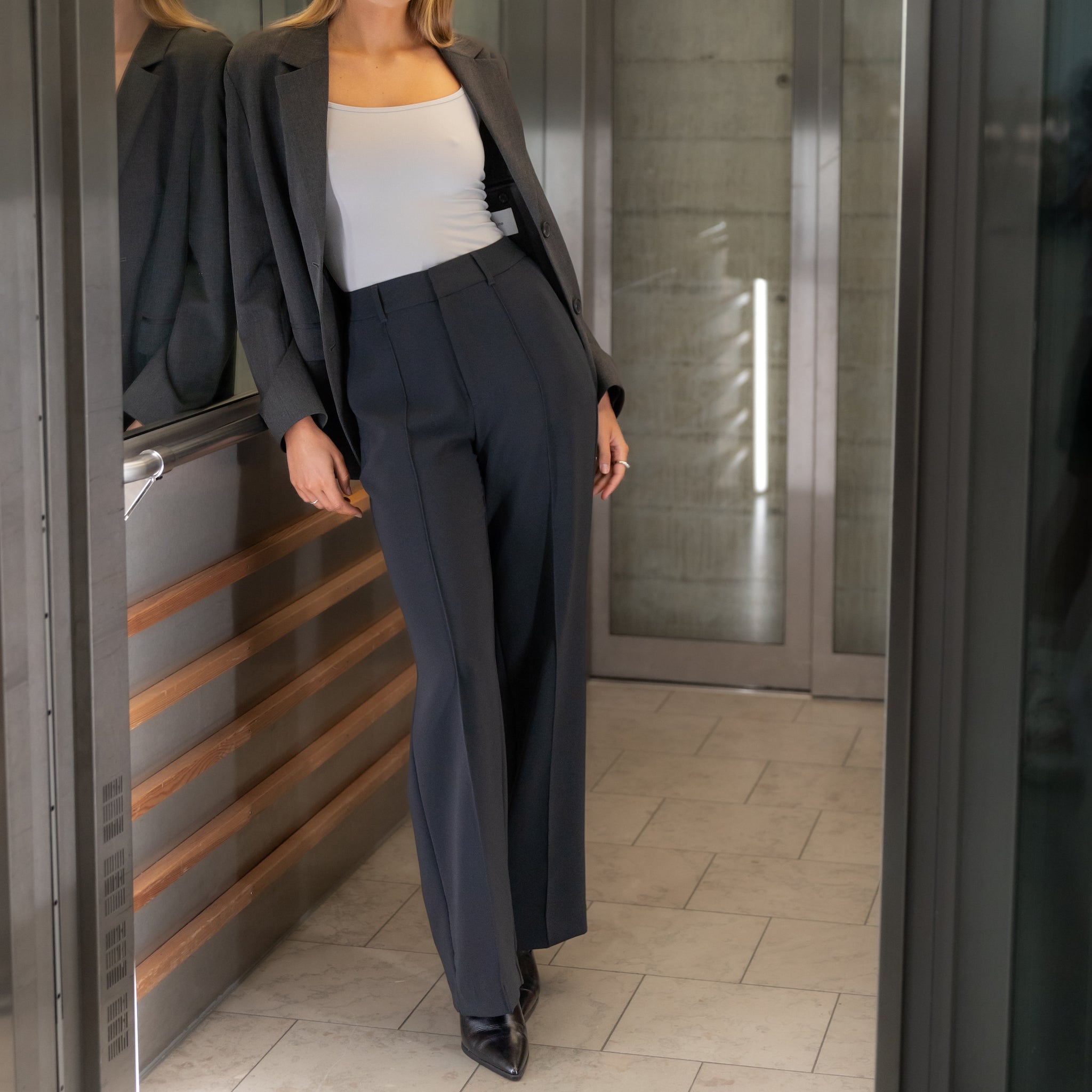 Model leaning against elevator railing wearing Ninepine Crepe Relaxed Trousers in Black with a white tank and grey blazer.