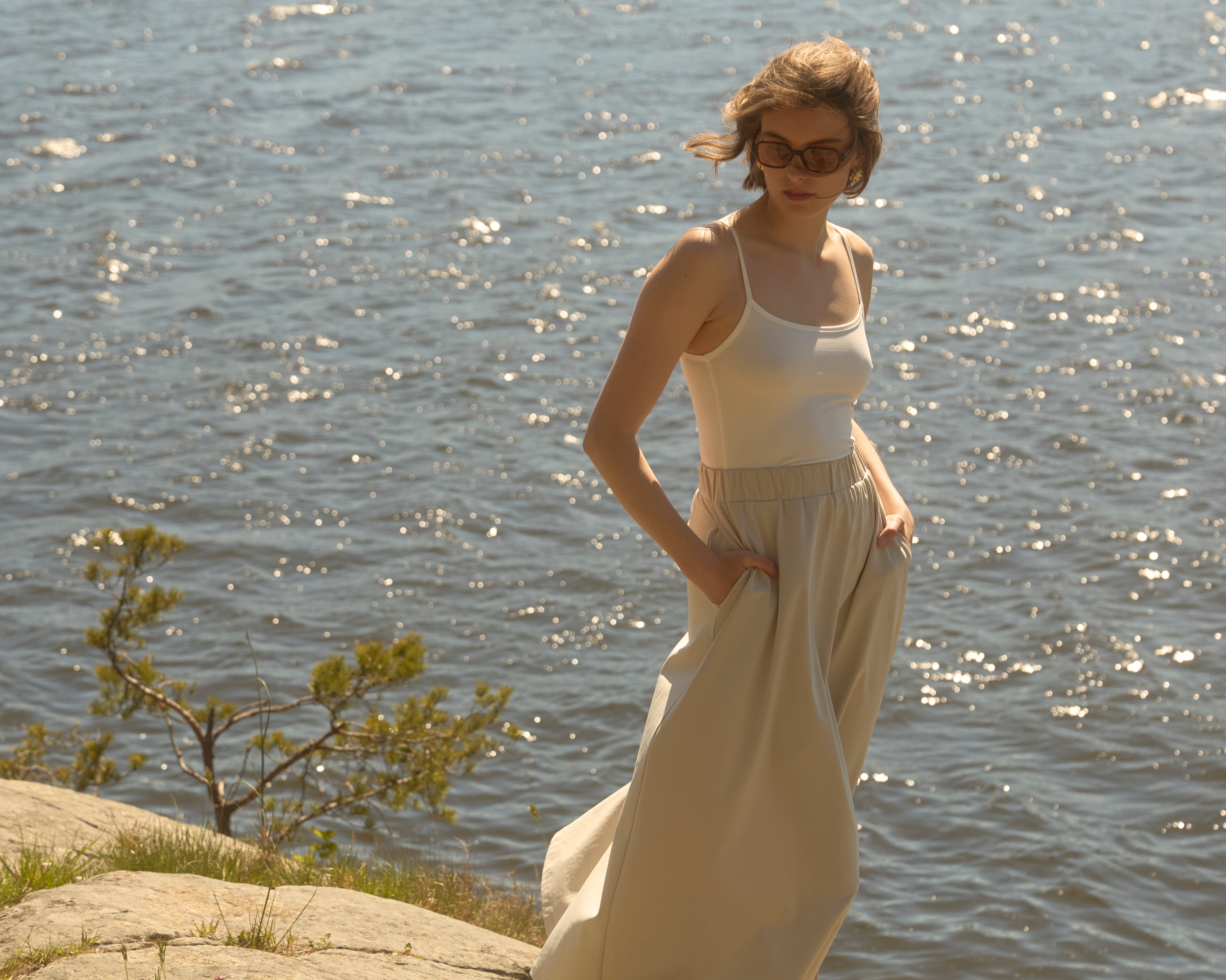 Wind-tousled woman by a sparkling Scandinavian coastline wearing Ninepine sand-beige flowy skirt and white tank, breezy summer look.