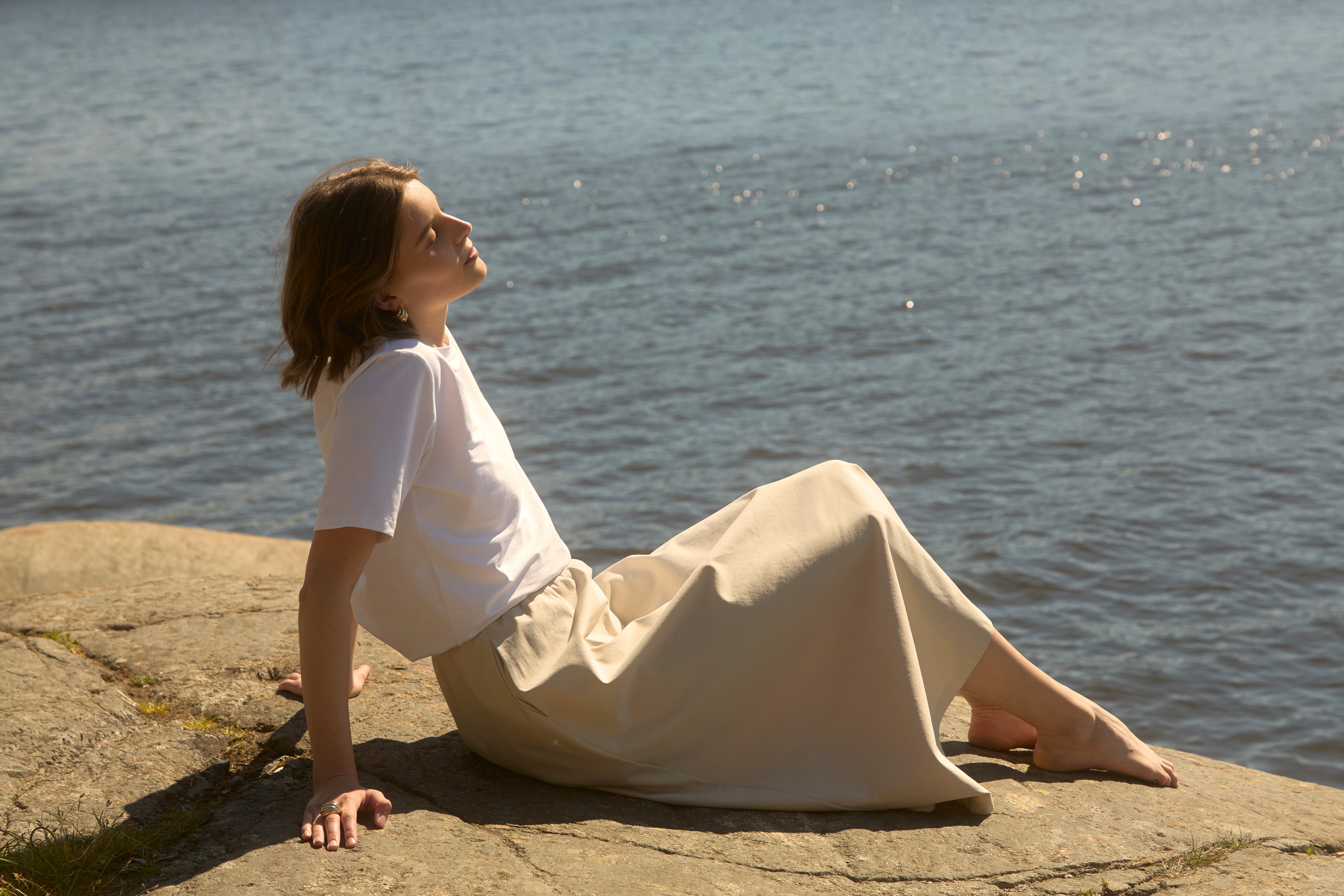 Woman in beige flowy Ninepine Flowy skirt and white tee reclining barefoot on a sun-warmed Nordic shoreline, serene summer minimalism