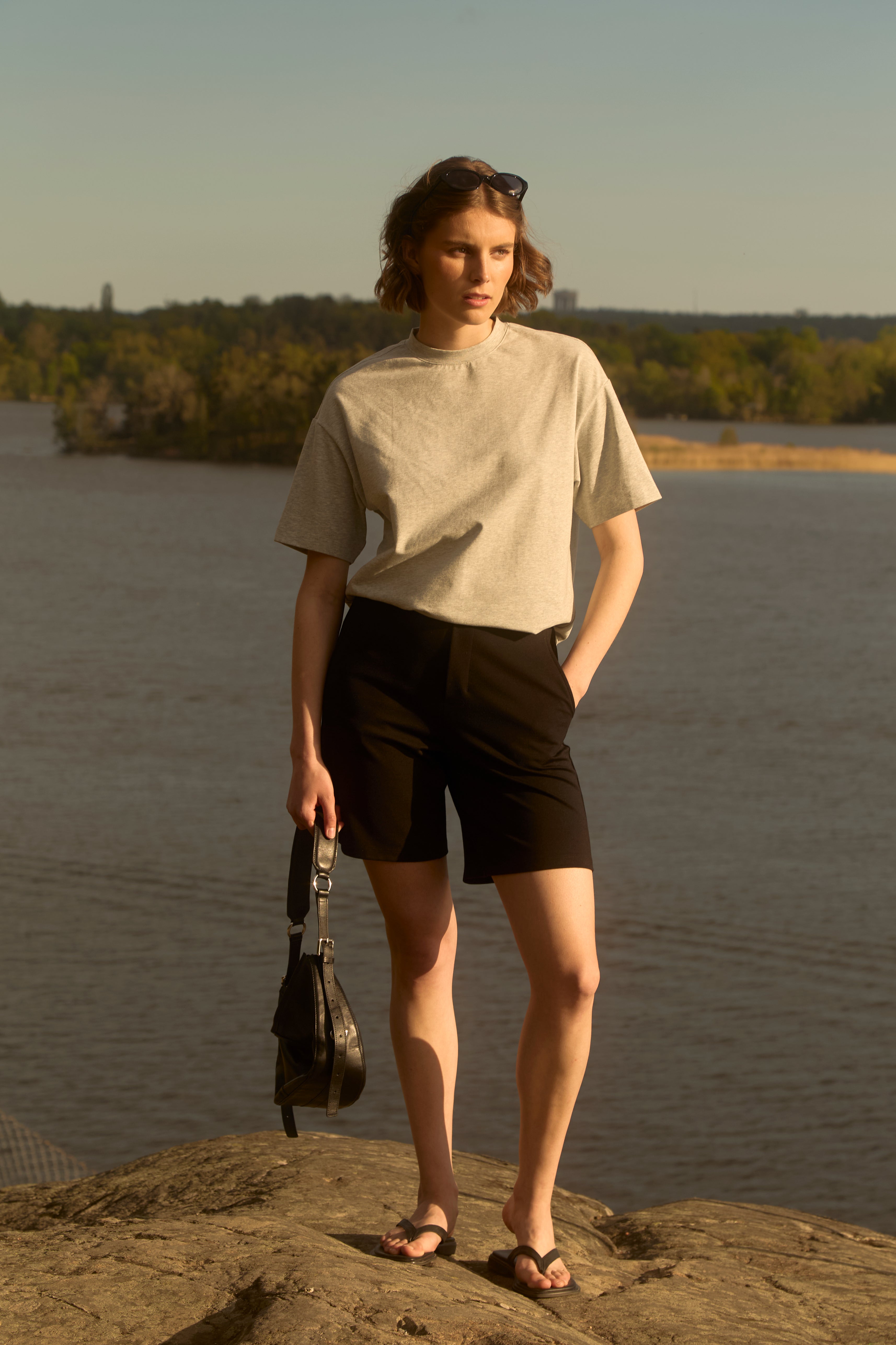Woman at golden-hour Swedish lakeshore wearing black Ninepine Light Ponte shorts and grey pima tee, holding a small bag—clean Nordic summer look.