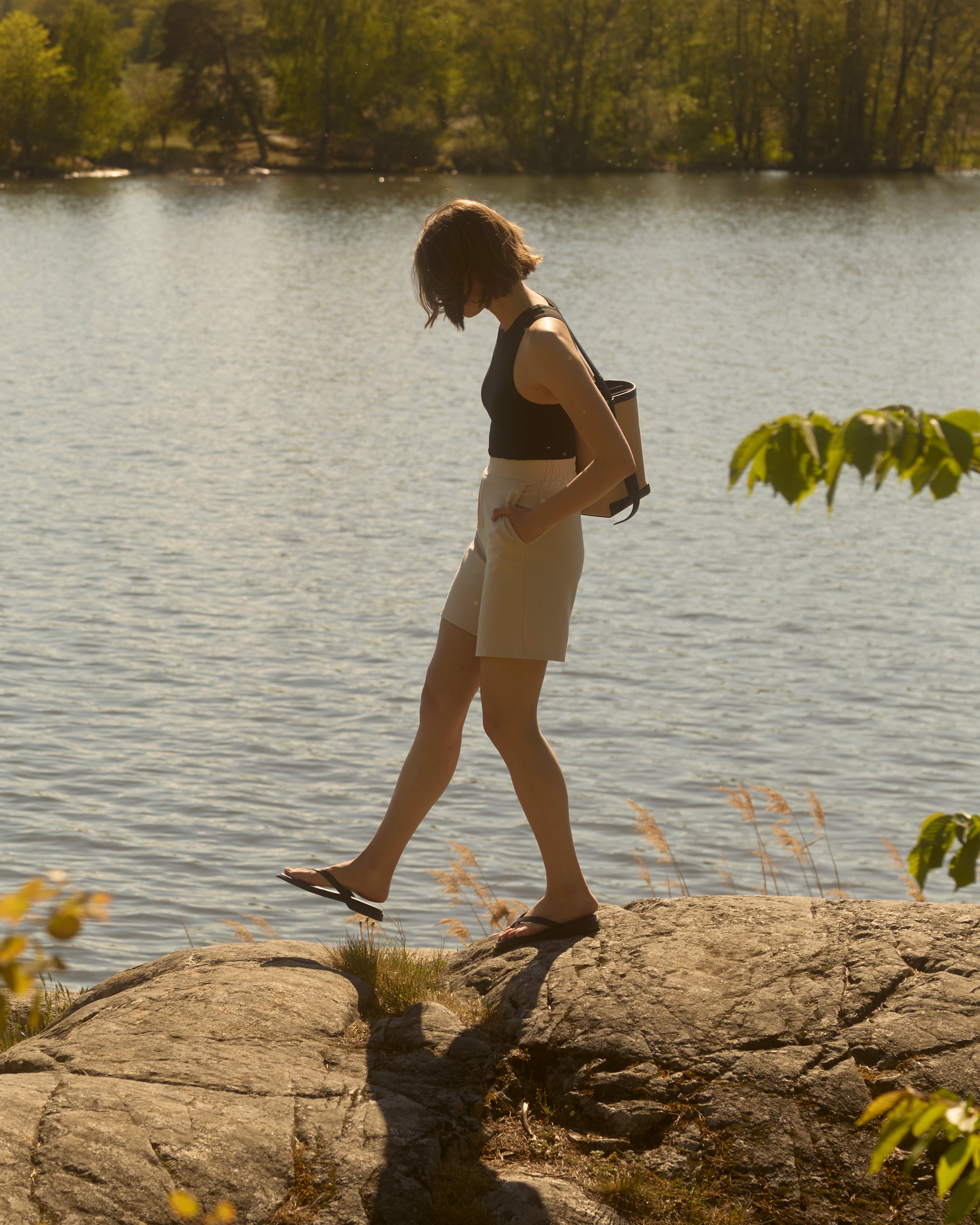 Side profile of woman walking on rocky lakeshore in sand-beige Ninepine Light Ponte shorts, black racerback tank and flip-flops, relaxed Swedish summer vibe.