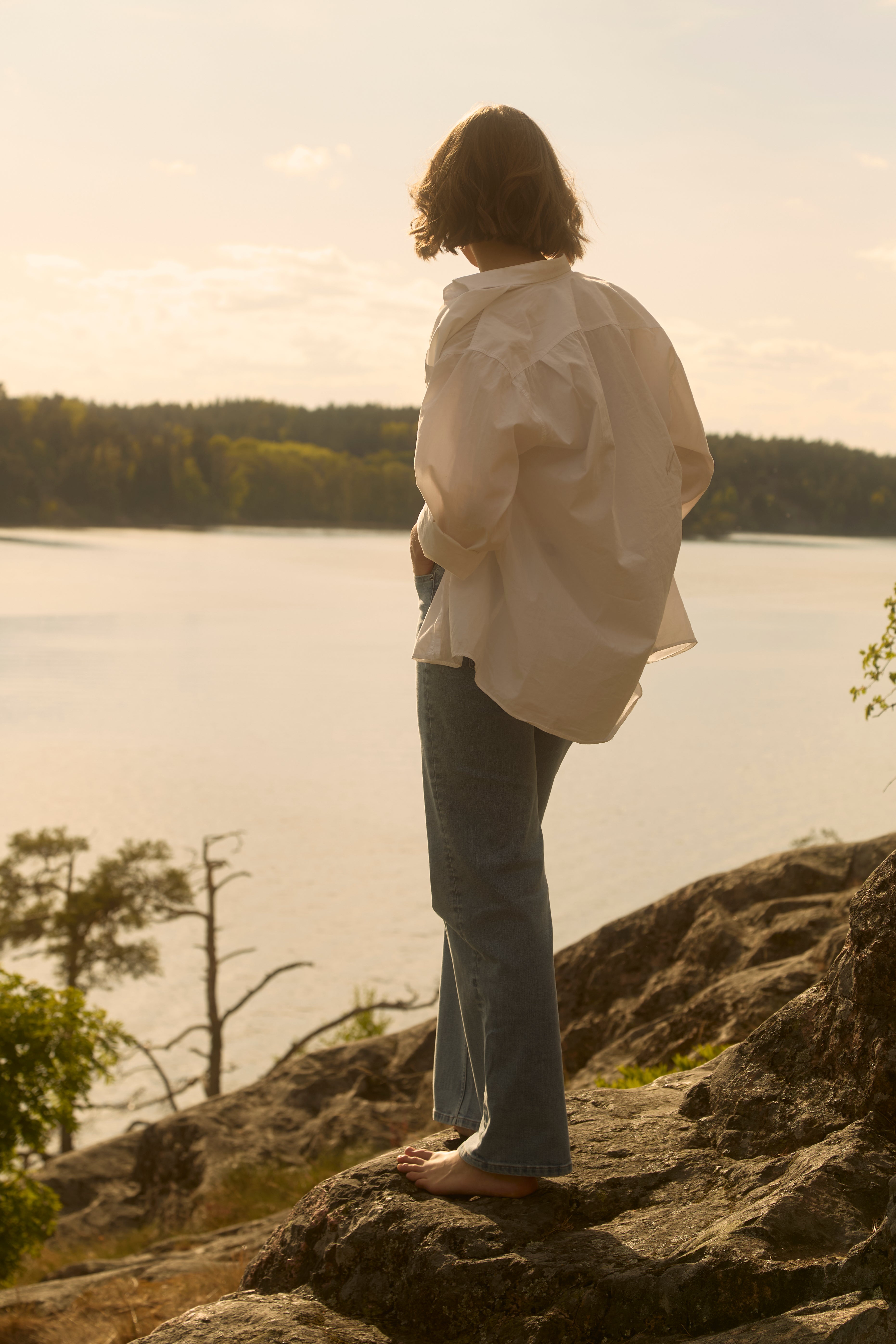 Barefoot woman in light-wash Ninepine Relaxed ComfortDenim jeans and billowy white shirt stands on a rocky Swedish lakeshore at golden-hour midsummer.