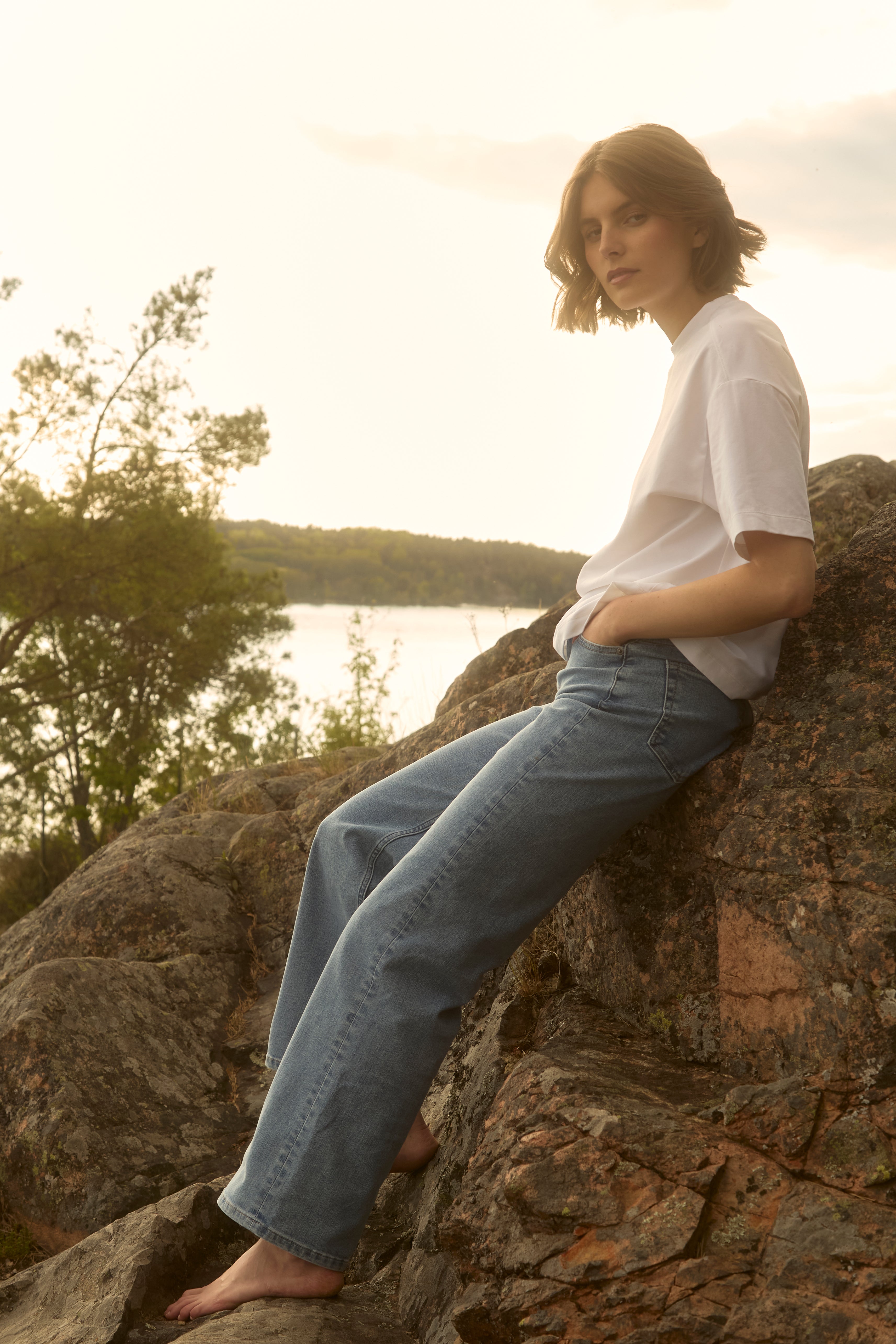 Barefoot woman in light-wash Ninepine relaxed ComfortDenim jeans and crisp white pima tee reclines on a sun-warmed Swedish lakeside rock at sunset, serene summer mood.